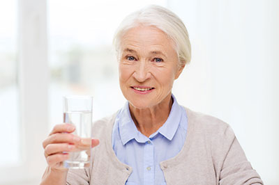 An elderly woman holding a glass of water, smiling at the camera with her hand raised slightly above the glass