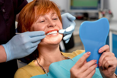 A woman sitting in a dental chair with a smile on her face, holding a blue mouthguard while a dentist adjusts it with tools