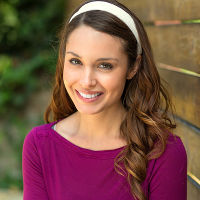 A young woman with long brown hair and a wide smile, posing against a wooden fence